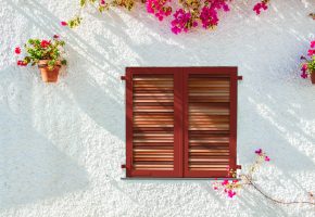 A blue wooden shuttered louvred window of a white washed spanish town house, with Bougainvillea and potted plants against the white washed wall.